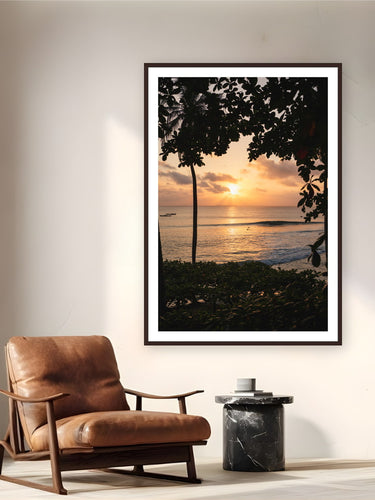 Black wood frame fine art print of Last Light by Ben Gamlin, Mentawai Islands, Sumatra, Indonesia, displayed on a reading nook wall — sunset in the Mentawais, palms in silhouette and a few surfers still in the water.