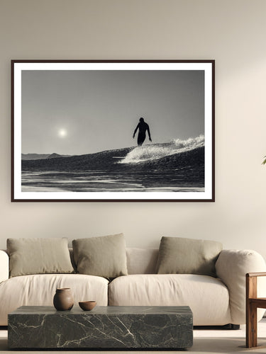 Black wood frame fine art print of Between Sun and Sea by Jeremy Bishop, Morro Bay, California, displayed on a modern living room wall — last light off Morro Bay — Jeremy's surfer silhouetted between the sun and the wave.