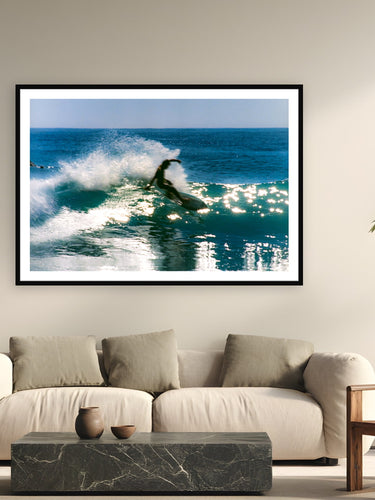 Black wood frame fine art print of Backlit Slide by Devon Cardamone, Malibu, California, displayed on a modern living room wall — late-afternoon Malibu, when the sun drops behind the lineup and punches straight through each wave as it feathers.