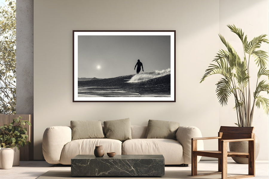 Black wood frame fine art print of Between Sun and Sea by Jeremy Bishop, Morro Bay, California, displayed on a modern living room wall — last light off Morro Bay — Jeremy's surfer silhouetted between the sun and the wave.