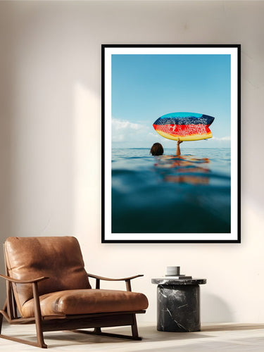 Black wood frame fine art print of Fishy by Sarah Swan, Mexico, displayed on a reading nook wall — between sets on a bigger swell day in Mexico — a rainbow fish board held overhead on a glassy sunrise morning.