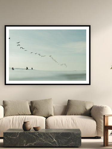 Black wood frame fine art print of Pelican Lineup by Sarah Swan, Costa Rica, displayed on a modern living room wall — a squadron of brown pelicans swept through the lineup in Costa Rica while three surfers waited for the last waves of the afternoon.