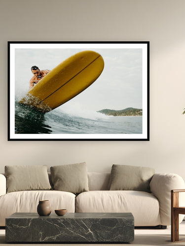 Black wood frame fine art print of Stinger by Sarah Swan, Mexico, displayed on a modern living room wall — a yellow longboard swinging straight at the camera during an afternoon free surf at the Mexi Log Fest in Mexico.
